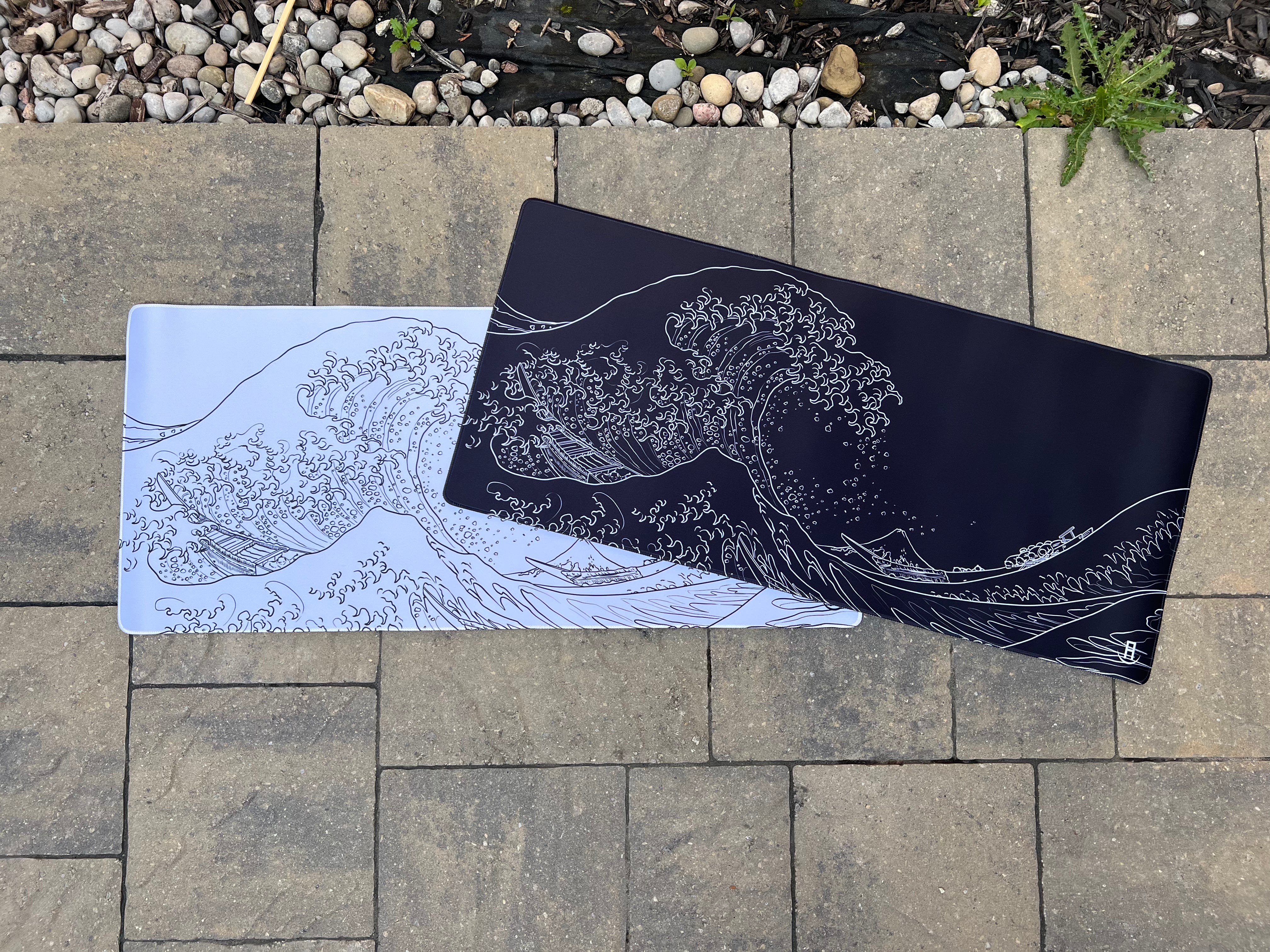 Two rectangular mousepads featuring a wave design inspired by Hokusai's 'The Great Wave off Kanagawa' are placed on a paved surface outdoors. One mousepad is white with a black wave design, while the other is black with a white wave design. Both are partially overlapping each other.