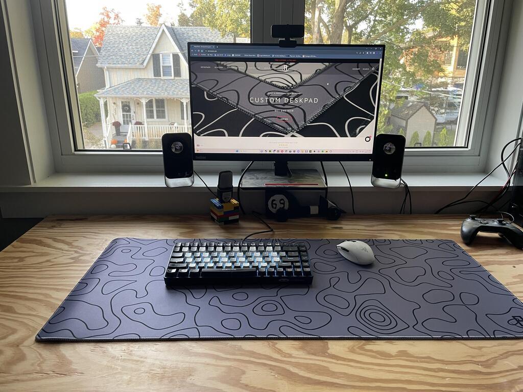 A home office setup with a monitor displaying a website on custom desk pads, placed on a wooden desk in front of a window. On the desk is a large grey mousepad with a topographic design, a mechanical keyboard with black and white keys, and a white wireless mouse. Two speakers flank the monitor, and a game controller is positioned on the right side of the desk.
