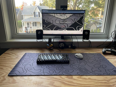 A home office setup with a monitor displaying a website on custom desk pads, placed on a wooden desk in front of a window. On the desk is a large grey mousepad with a topographic design, a mechanical keyboard with black and white keys, and a white wireless mouse. Two speakers flank the monitor, and a game controller is positioned on the right side of the desk.