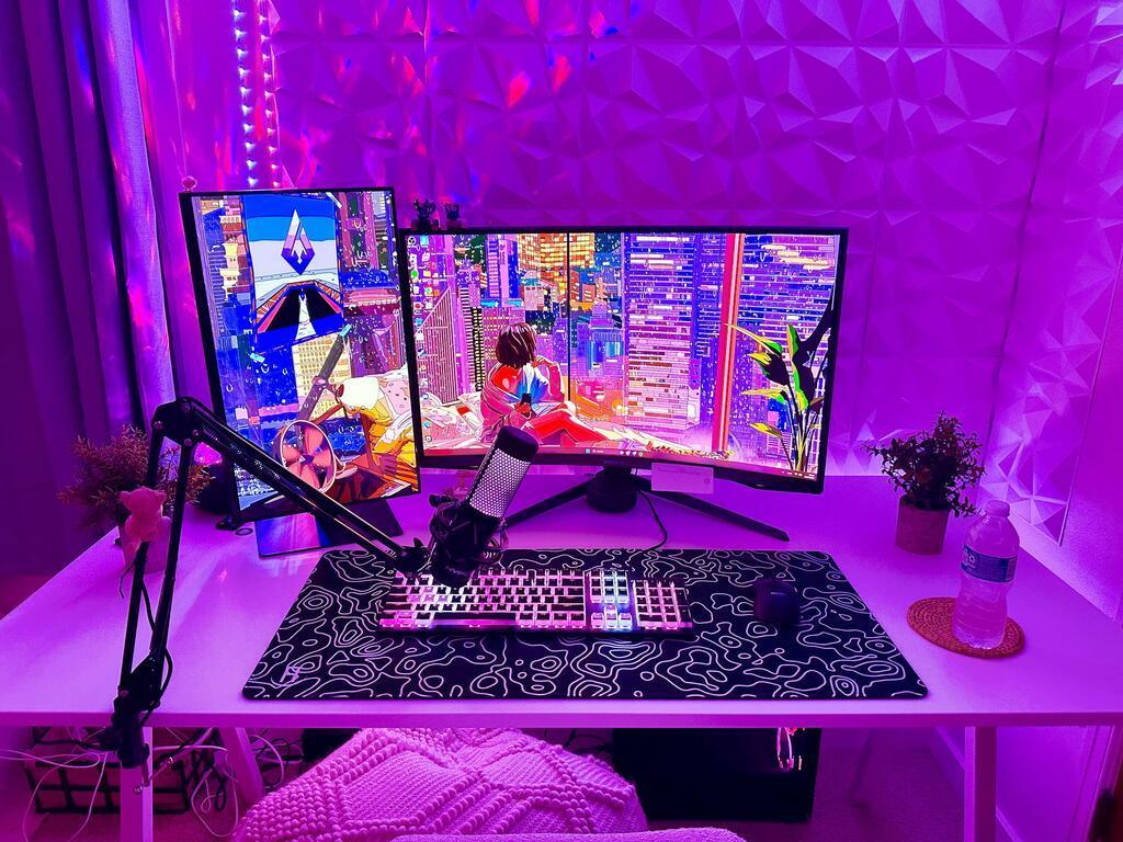 A computer setup with two monitors, a mechanical keyboard, a microphone on a boom arm, and a water bottle on a desk. The room is lit with pink and purple LED lights, Black topography mousepad and deskpad on gaming desk