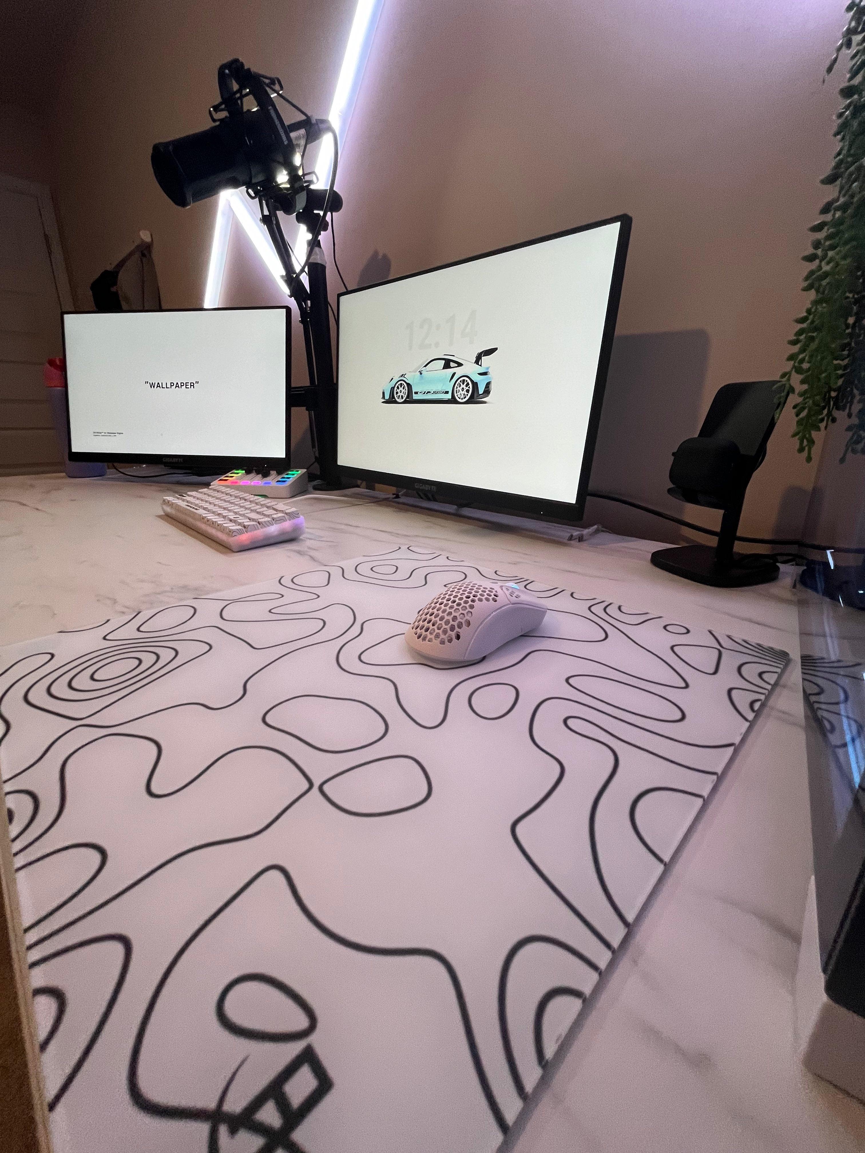 A stylish computer setup featuring two monitors, a white mechanical keyboard, a white gaming mouse with a honeycomb design, and a large glass mousepad with abstract black line patterns on a white background. The desk has a marble-like surface, and there is a microphone mounted on an adjustable arm. The setup is illuminated by diagonal LED strip lights on the wall, adding a modern and vibrant touch.