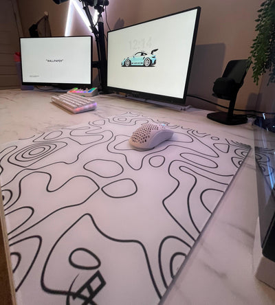 A stylish computer setup featuring two monitors, a white mechanical keyboard, a white gaming mouse with a honeycomb design, and a large glass mousepad with abstract black line patterns on a white background. The desk has a marble-like surface, and there is a microphone mounted on an adjustable arm. The setup is illuminated by diagonal LED strip lights on the wall, adding a modern and vibrant touch.