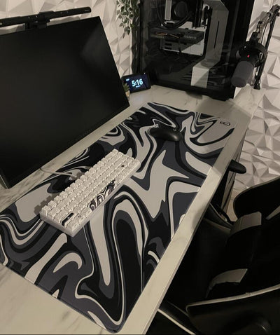 A sleek gaming setup featuring a black monitor, a black and white mechanical keyboard, and a black wireless mouse on a large mousepad with a black, white, and grey marble pattern. The setup is on a white desk with a geometric white wall in the background. To the right is a gaming PC with visible components through a transparent side panel. An overhead lamp, a microphone on an arm, and a digital clock displaying '5:16' complete the setup.
