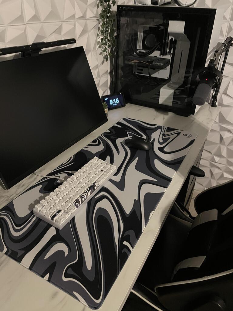 A sleek gaming setup featuring a black monitor, a black and white mechanical keyboard, and a black wireless mouse on a large mousepad with a black, white, and grey marble pattern. The setup is on a white desk with a geometric white wall in the background. To the right is a gaming PC with visible components through a transparent side panel. An overhead lamp, a microphone on an arm, and a digital clock displaying '5:16' complete the setup.